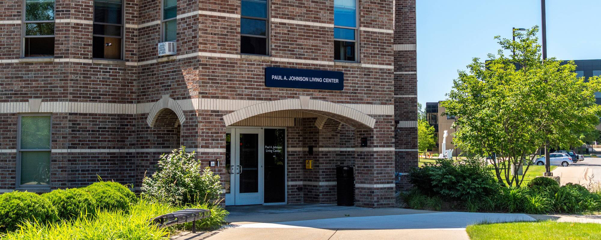 Red-brick building corner with an entrance under an arch. A sign above reads "Paul A. Johnson Living Center." Lush green shrubs and trees surround the area.